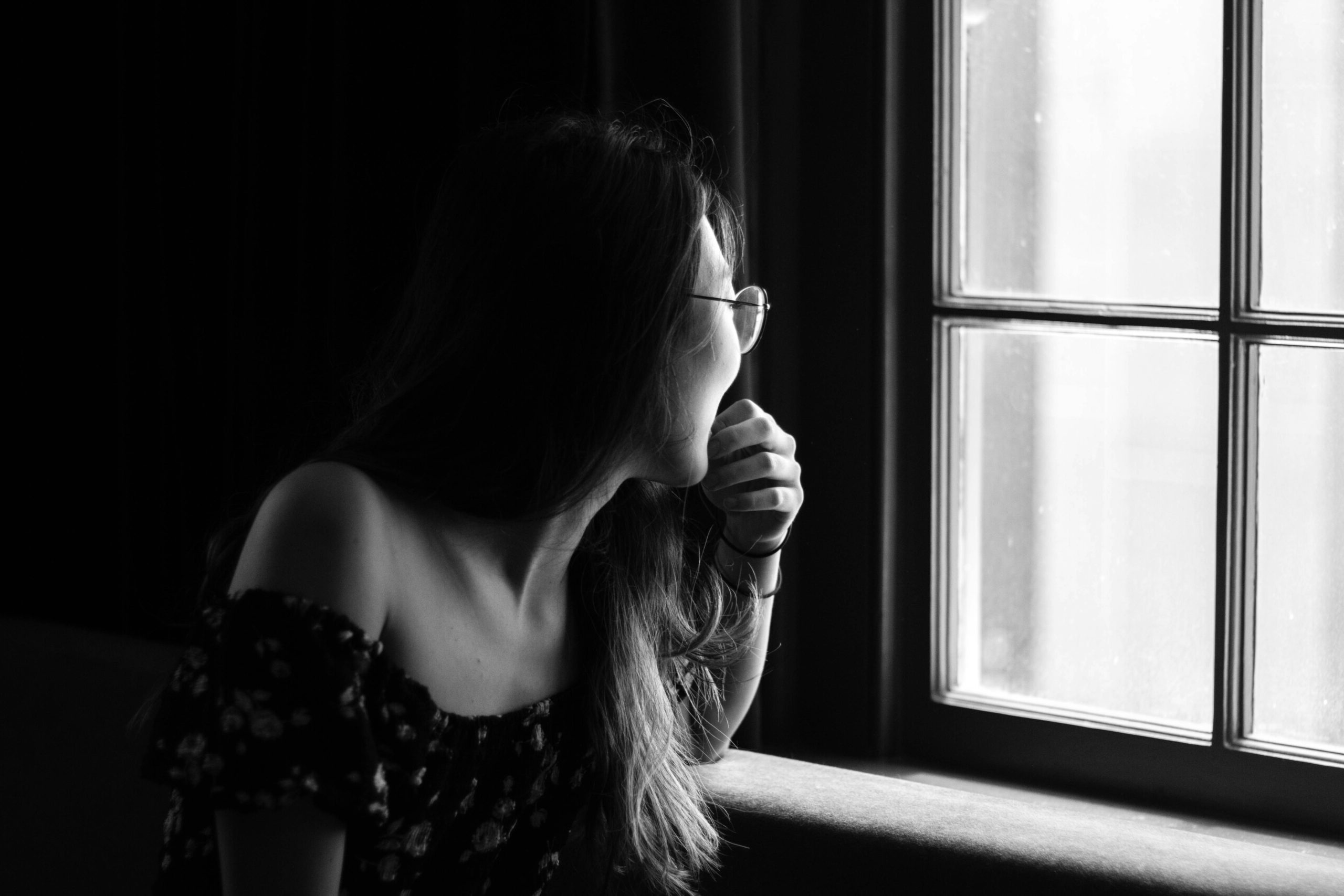 Grayscale photo of a young woman staring out of a window. The room she is in is dark, while there is light coming in through the window. She appears to be wearing glasses. There is nothing visible outside the window.